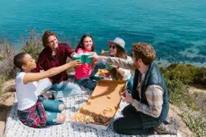 Fünf Freunde auf einer Parkdecke am Meer, sich zuprostend mit Pizza
